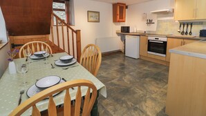 A compact kitchen with wooden cabinets, a dining table set for four, and a small refrigerator. - Graig Las The Stables (Oswestry)