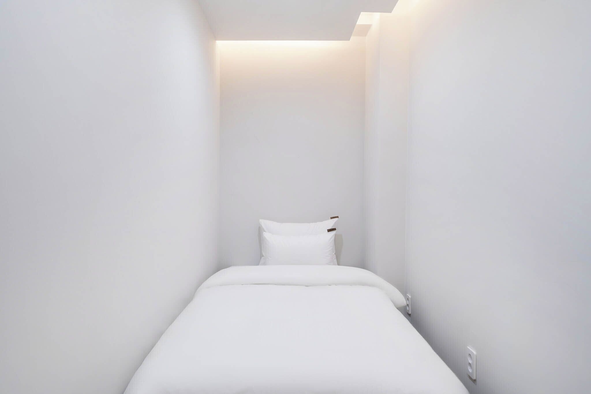 A narrow, white-walled room with a single bed and a small bedside table.