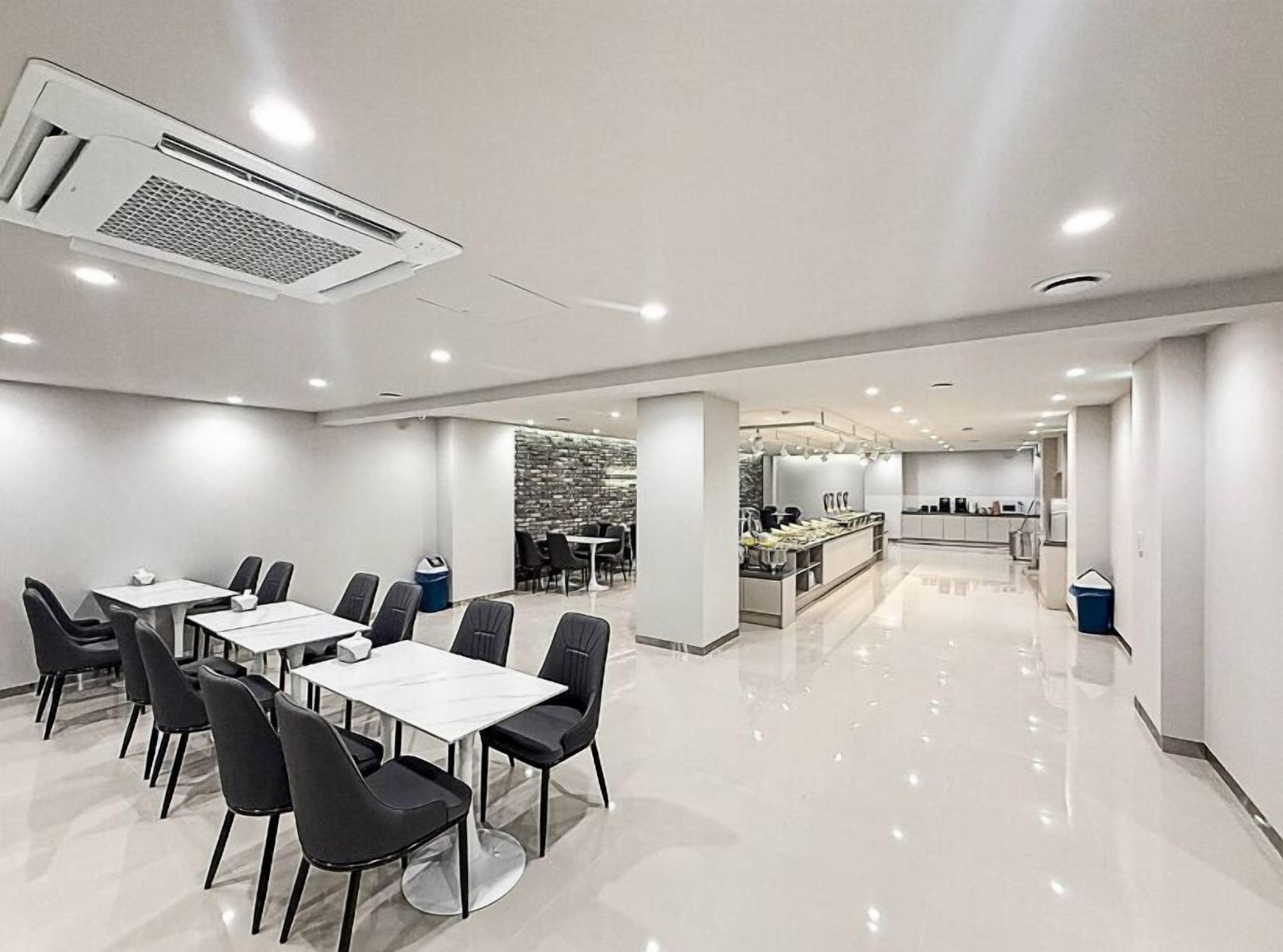 Un comedor de hotel moderno con mesas blancas y sillas negras, una cocina al fondo y un revestimiento de pared de piedra.