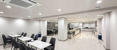 A modern hotel dining area with white tables and black chairs, a kitchen in the background, and a stone wall feature.