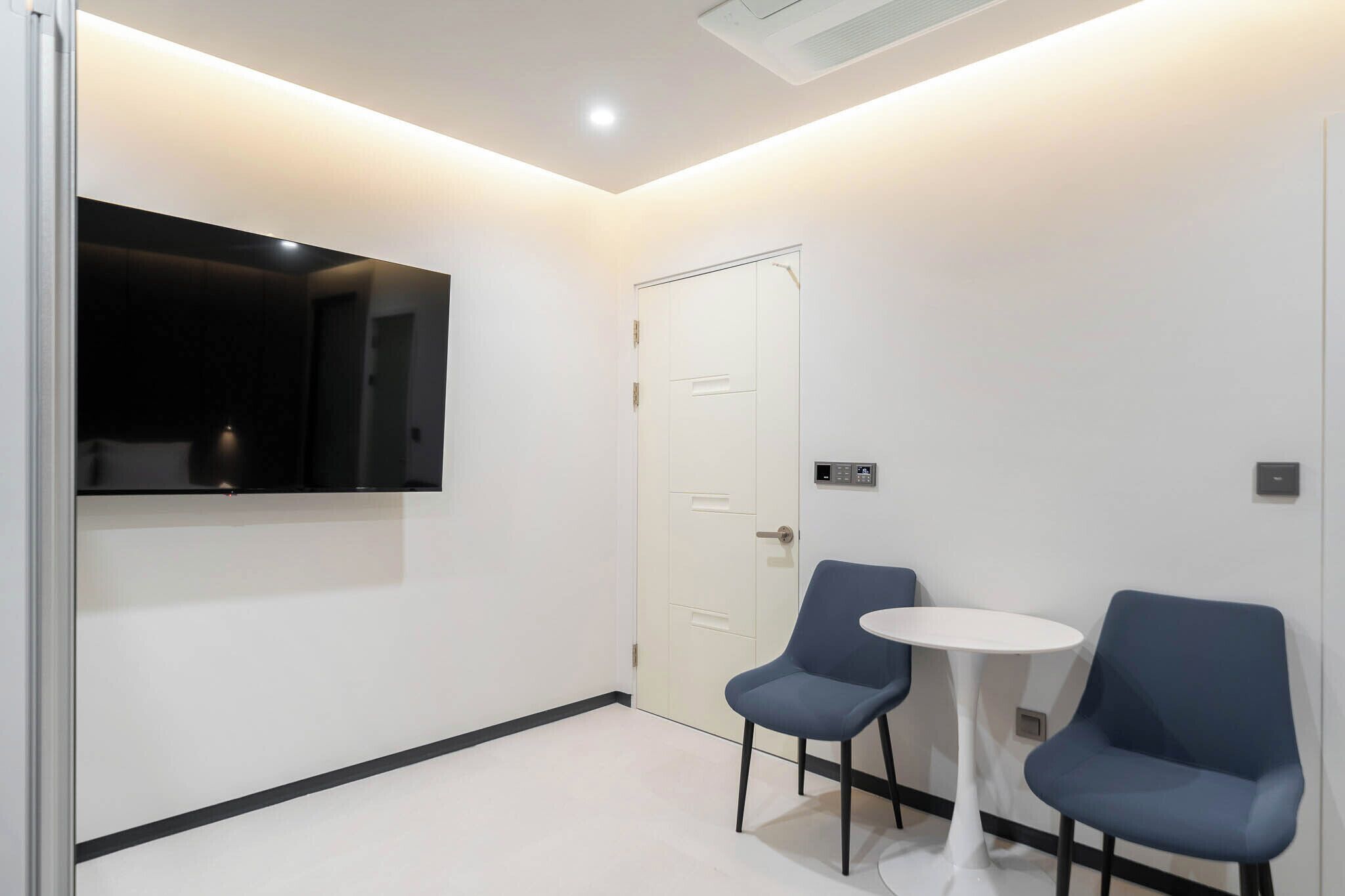 Una habitación moderna con una puerta blanca, un televisor de pantalla plana montado en la pared y dos sillas azules alrededor de una pequeña mesa blanca.