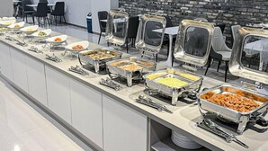 Un buffet est installé avec divers plats présentés dans des récipients en métal, disposés sur un long comptoir. On aperçoit en arrière-plan des chaises et des tables.