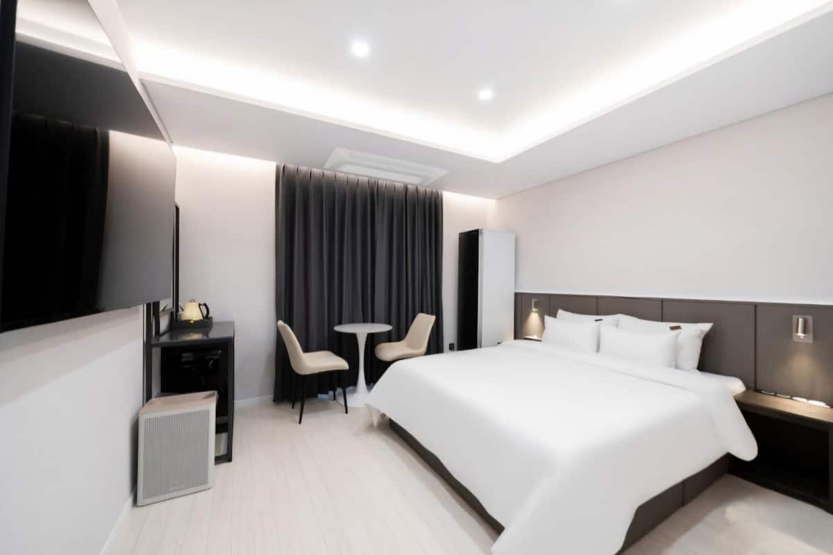 Ein modernes Hotelzimmer mit einem großen Bett, einem Schreibtisch mit Stuhl, einem kleinen Tisch für zwei Personen, einem Fernseher und einer Klimaanlage.