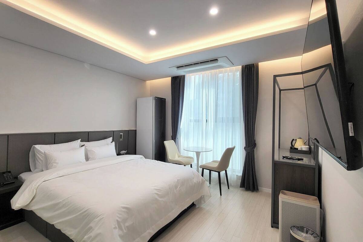 Ein modernes Hotelzimmer mit einem großen Bett, Nachttischen, einem Schreibtisch mit Stuhl, einem Flachbildfernseher und einem fenster mit Vorhängen.