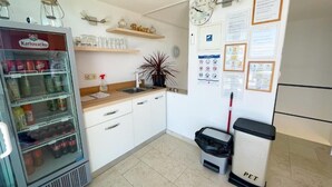 Ett litet pentry med kylskåp, diskho och en papperskorg. Det finns hyllor med glas och en klocka på väggen.
