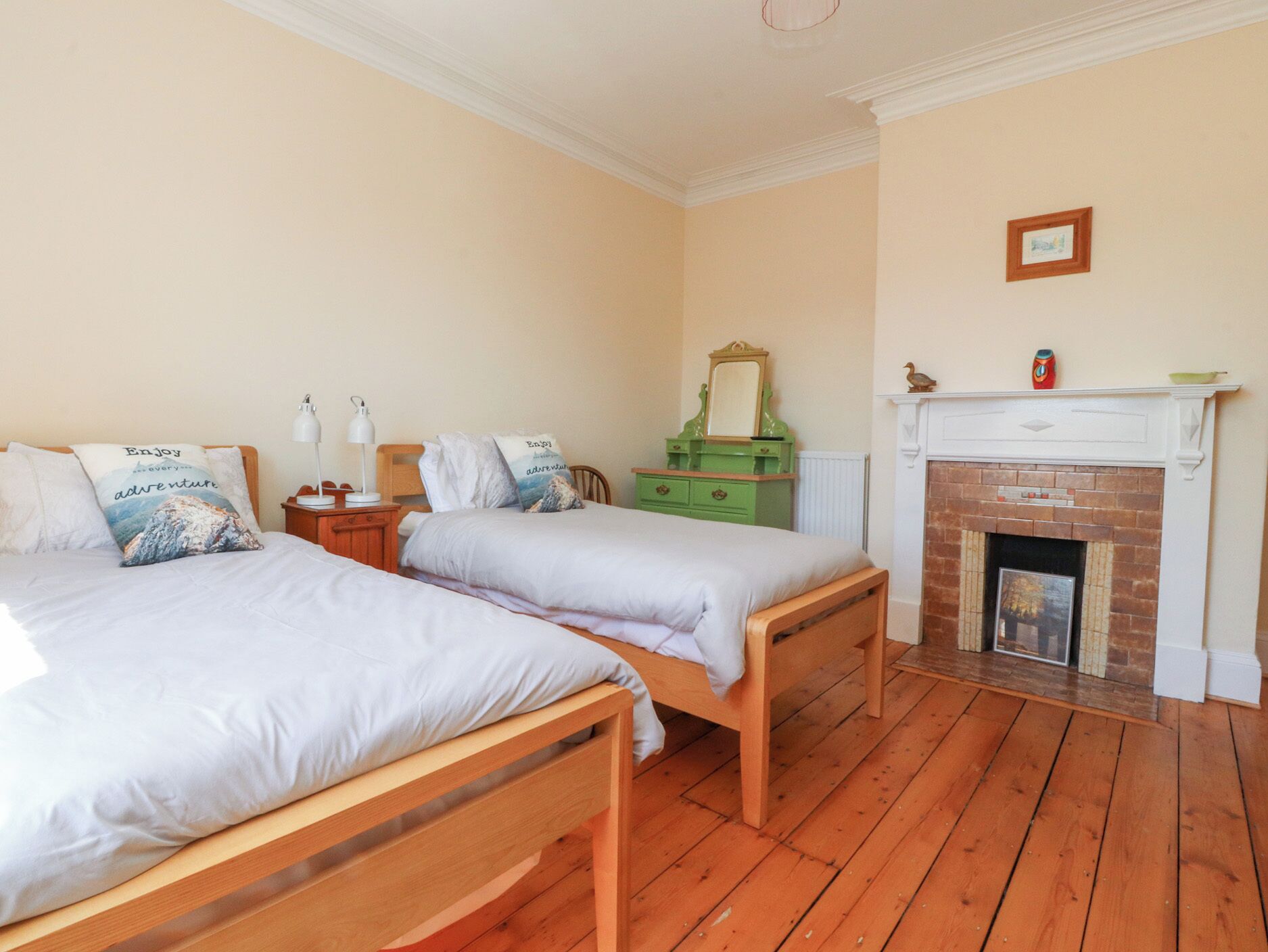 A bedroom with two wooden beds, a green dresser, a white fireplace, and a small framed picture on the wall.