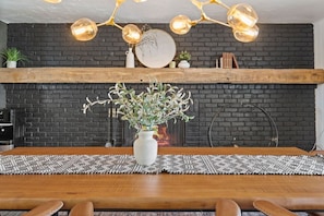 Een eethoek met een houten tafel, een witte vaas met groen, een zwarte bakstenen muur en een moderne kroonluchter.