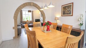 A dining area with a wooden table and chairs, a vase of red flowers, and a stone archway leading to a living room. - Cherry Tree Cottage (Bakewell)
