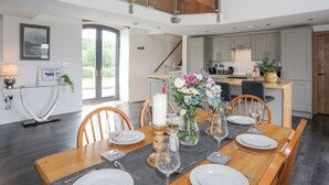 A dining area with a wooden table set for four, a vase with flowers, and a view of the kitchen and a staircase. - Gwel Y Fenai (Caernarfon)