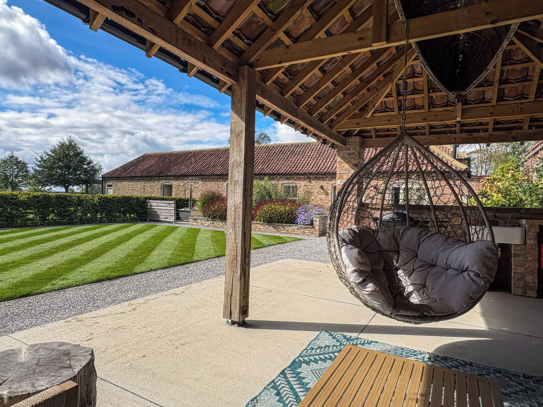 A covered outdoor seating area with a hanging chair, a wooden table, and a patterned rug.