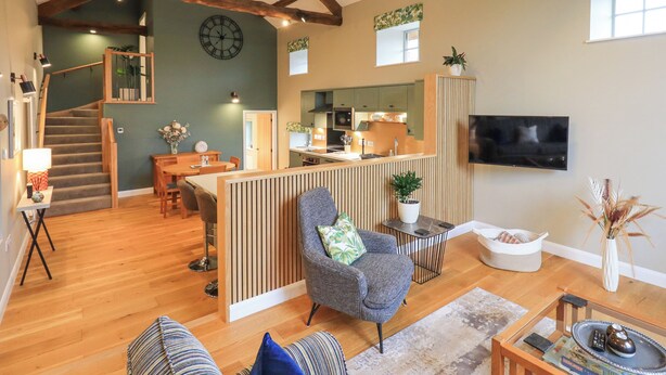 A spacious living area with a wooden staircase, a kitchen with a modern design, a comfortable seating area with a sofa and armchairs, and a flat-screen TV mounted on the wall. - Old Howgate Barn (Ambleside)