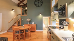 A modern kitchen with a wooden dining table, a clock on the wall, and a staircase leading to another level. - Old Howgate Barn (Ambleside)