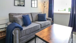Un soggiorno moderno con un divano grigio, due cuscini blu, un tavolino in legno e un'ampia finestra con tende azzurre.