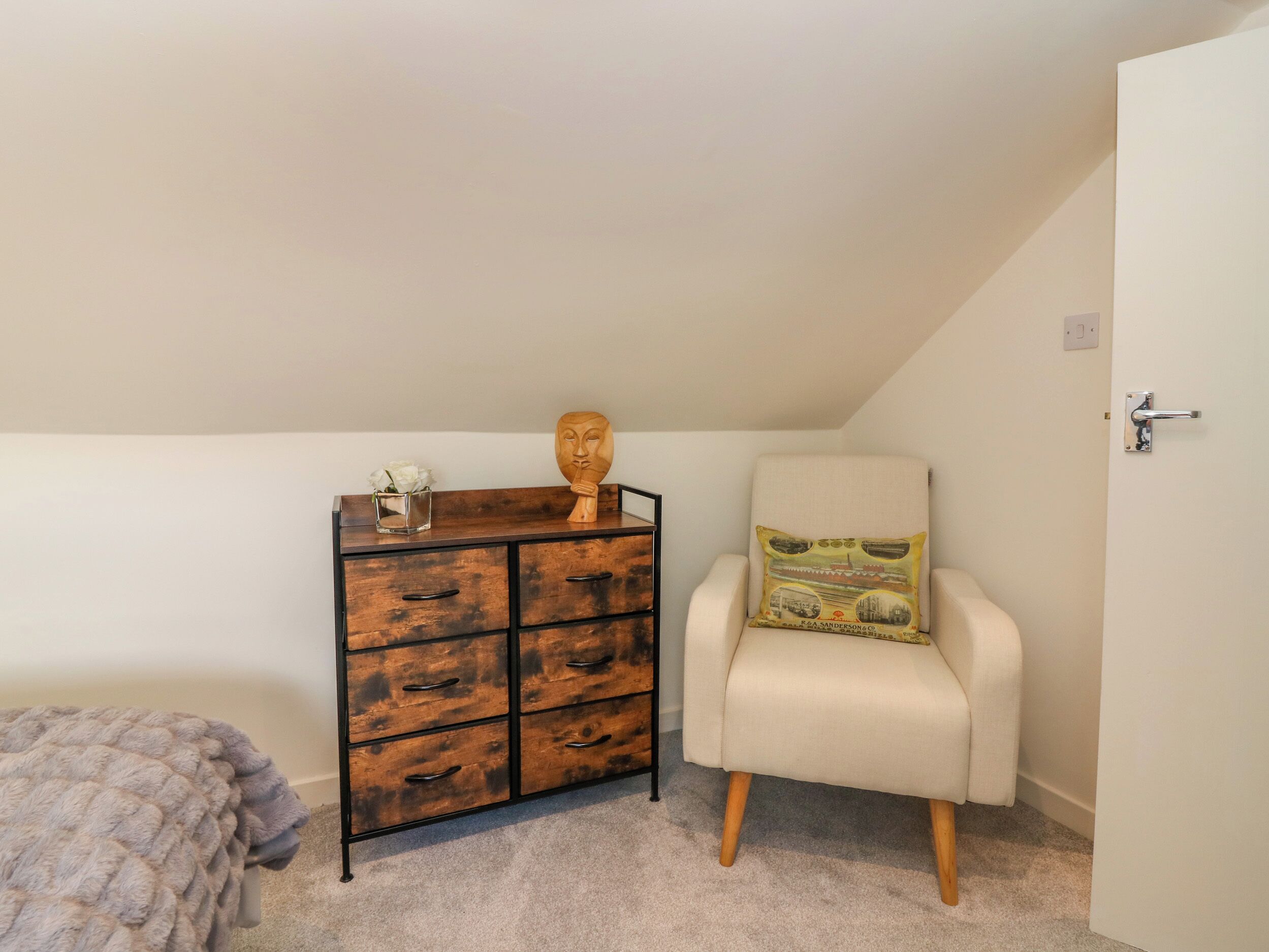 A bedroom with a wooden dresser, a white armchair, and a small table with a vase of flowers.