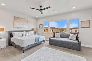 A bedroom with a bed, a gray sofa, large windows, and a ceiling fan.