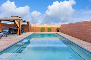 A rectangular swimming pool with a tiled deck, a covered seating area with chairs, and a brick wall.