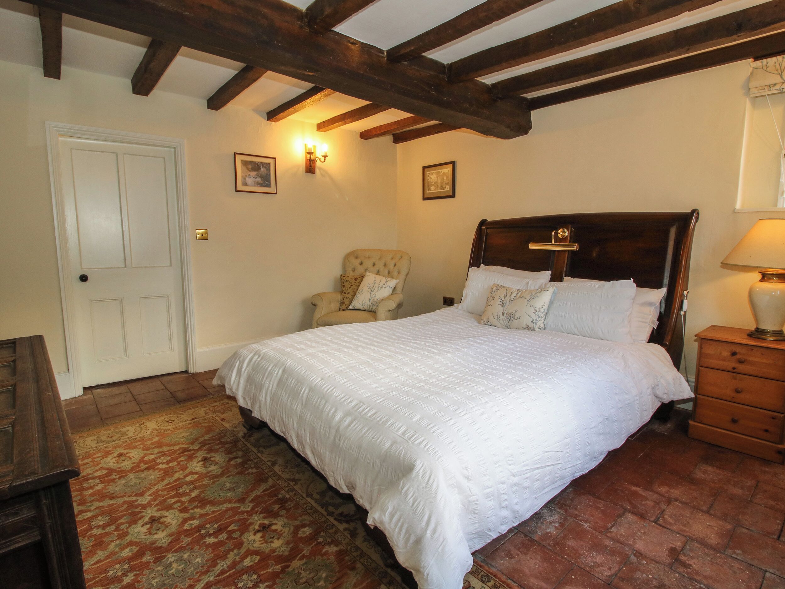 A bedroom with a large bed, a chair, a wooden nightstand, a lamp, and a door.