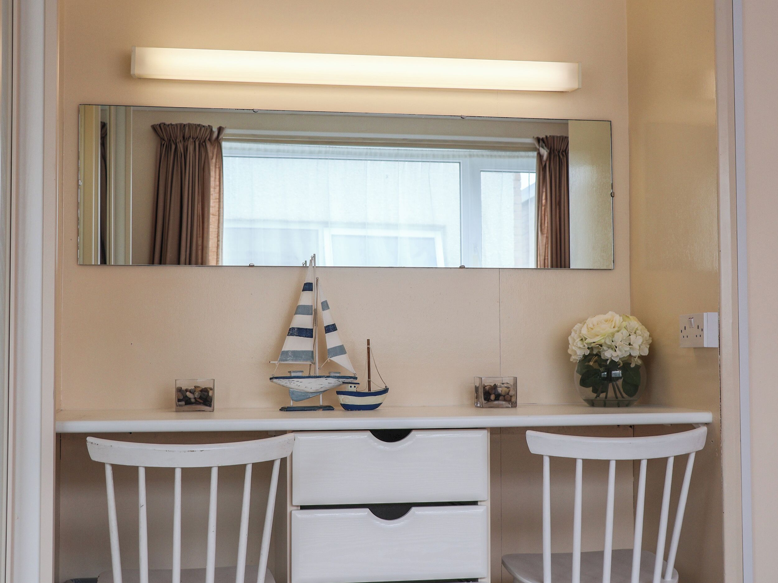 A room with a white desk, two white chairs, a mirror, a small sailboat model, a vase with flowers, and a window with curtains.