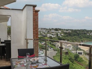 Ein Balkon mit Korbmöbeln, einem Glastisch und Blick auf eine Hügelstadt und einen Strand.
