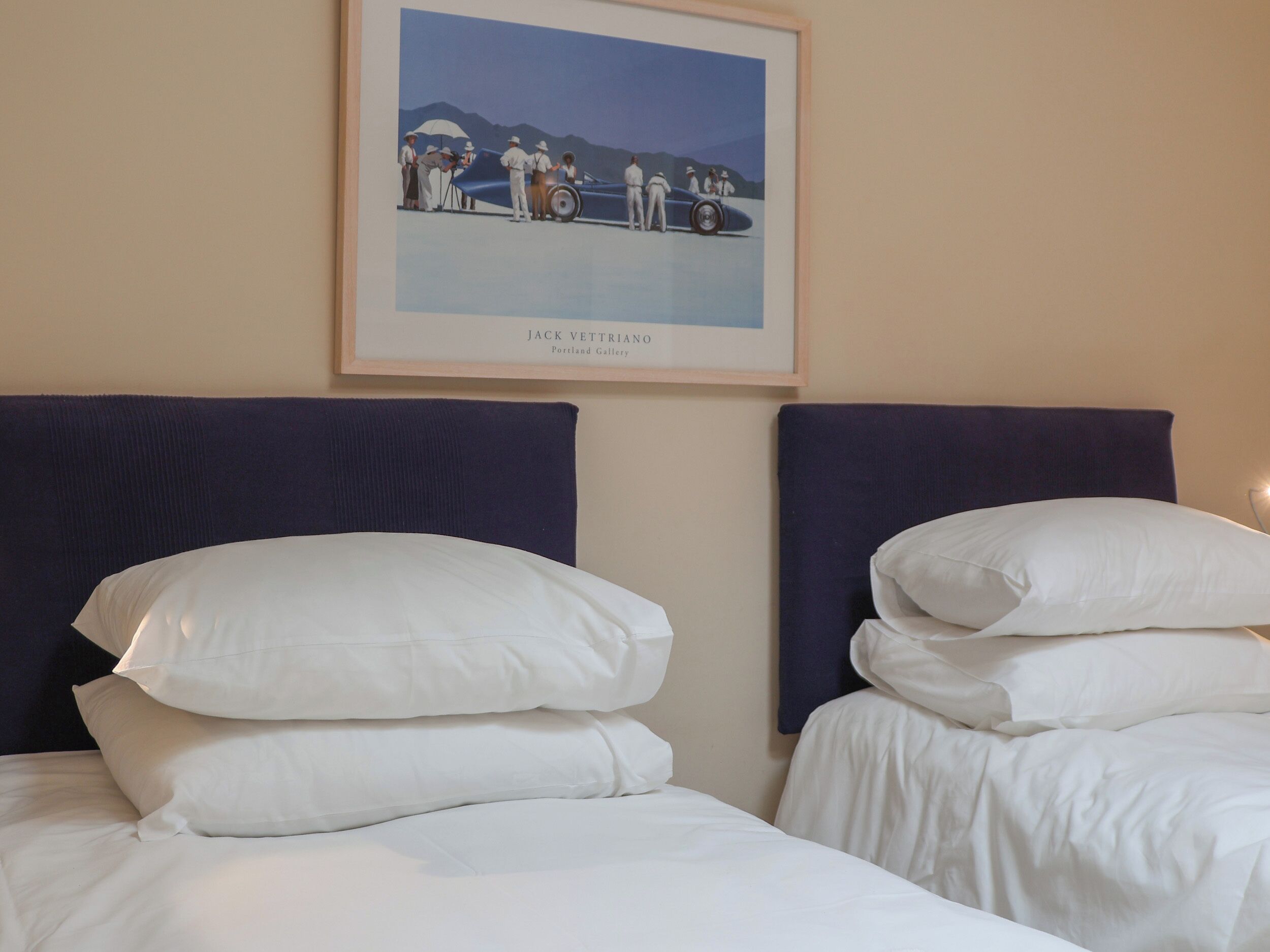 Camera d'albergo con due letti, ognuno con biancheria da letto bianca e testiere scure. Un quadro raffigura un gruppo di persone vicino a un'auto ed è appeso al muro sopra i letti.