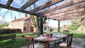 An outdoor patio with a wooden table set for dining, surrounded by wooden chairs with cushioned seats. - Woodpit Cottage (Nottingham)