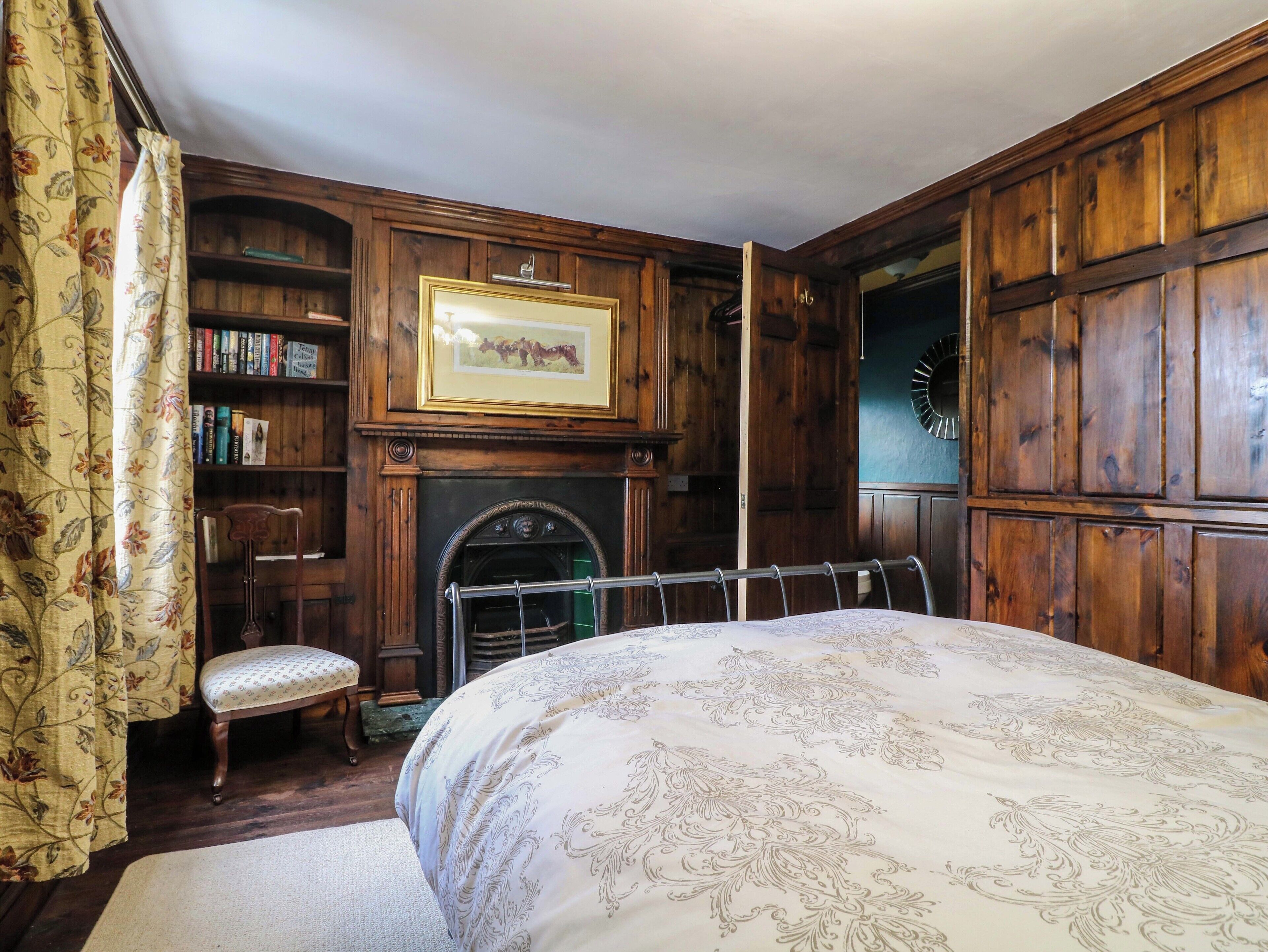 A bedroom with a large bed, wooden walls, a fireplace, a bookshelf, and a floral curtain.