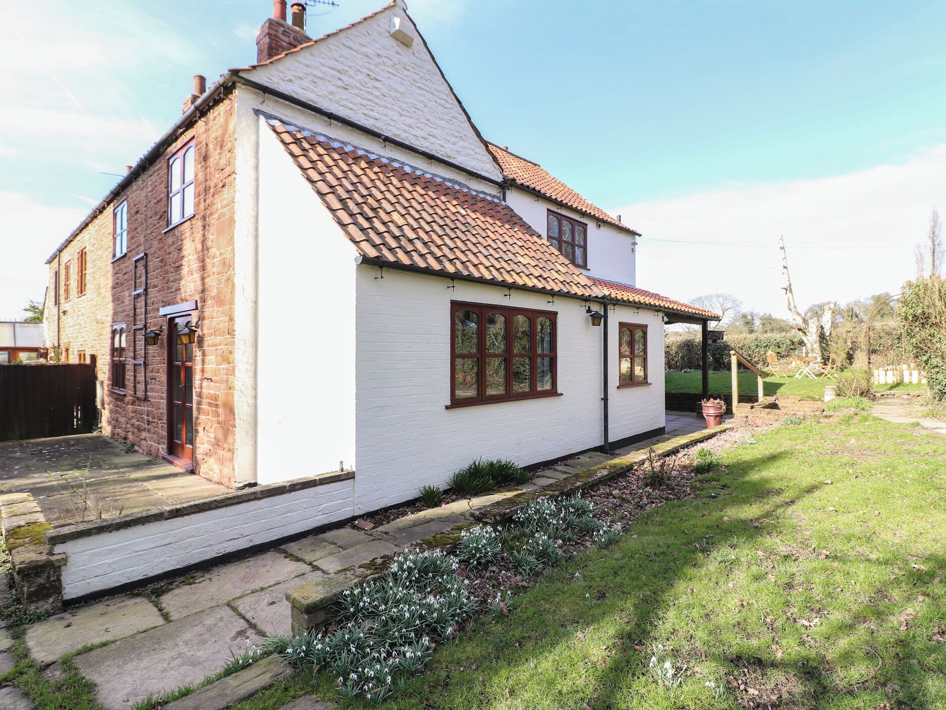 A white house with a tiled roof, a stone foundation, and a small garden with snowdrops.