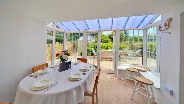 A dining area with a round table set for four, a glass-enclosed patio, and a view of the garden. - Ty Ffynnon (Pwllheli)