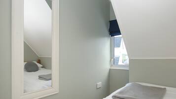 Un dormitorio con techo inclinado, un espejo grande, una cama con manta gris y un ventanal con persiana oscura.