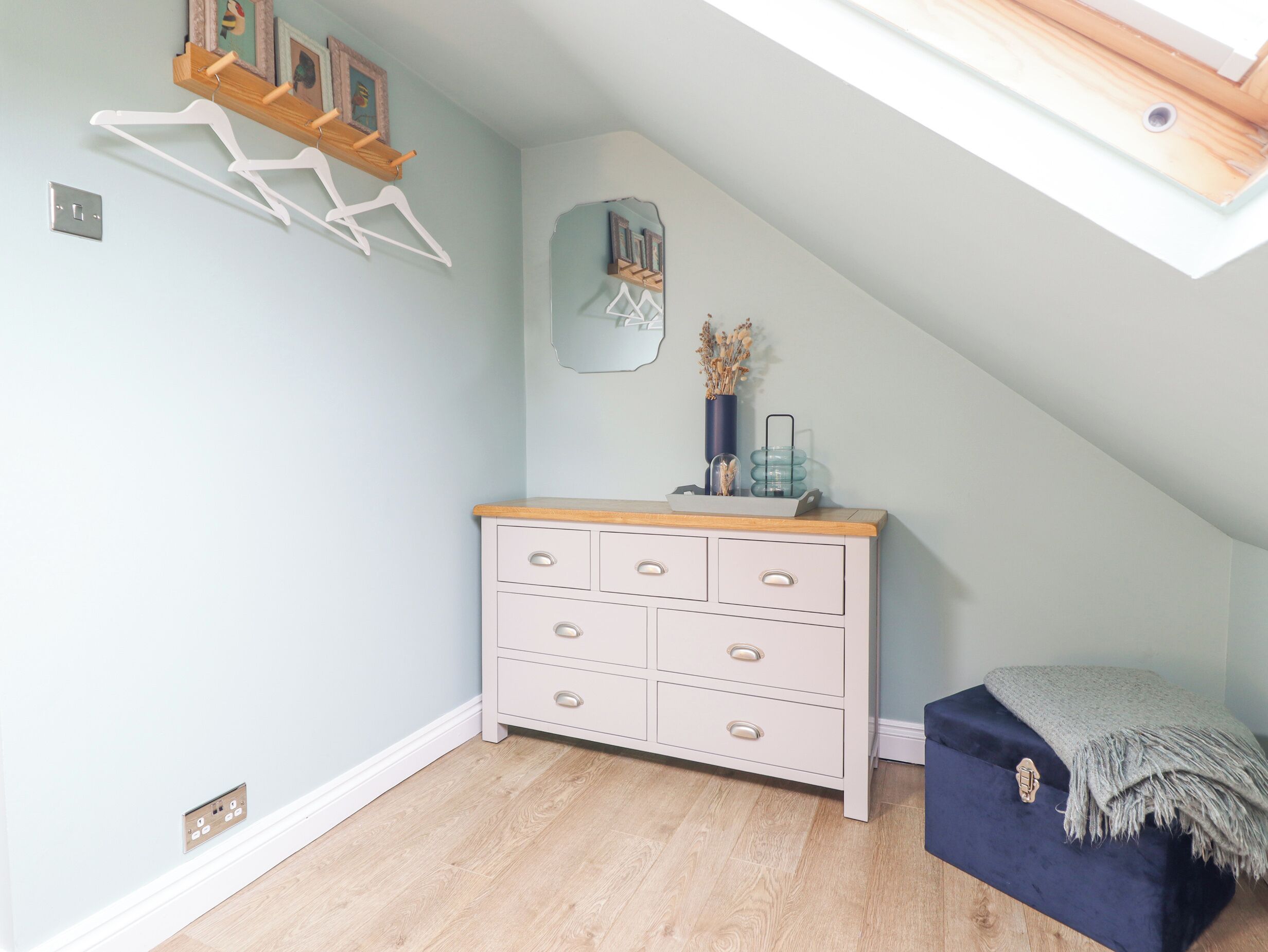 A room with a sloped ceiling, a white dresser with drawers, a blue bench, and a wooden shelf with pictures.