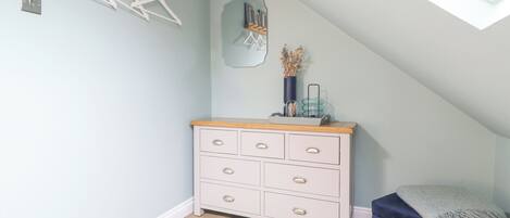 A room with a sloped ceiling, a white dresser with drawers, a blue bench, and a wooden shelf with pictures.