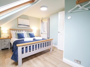 Ein Schlafzimmer mit schrägen Wänden, einem Bett mit Holzrahmen, einem Nachttisch mit Lampe und einem Fenster mit Vorhängen.