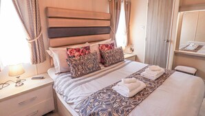 Ein ordentlich bezogenes Bett mit einem gemusterten Bettüberwurf, Kissen und gefalteten Handtüchern. Ein Nachttisch mit Lampe und Fernbedienung steht daneben. Das Zimmer verfügt über ein Holz-Kopfstück, Vorhänge und einen Spiegel an der Wand.