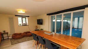 A spacious room with a wooden dining table, black chairs, a bean bag, a radiator, a TV, and a view of a swimming pool through a sliding glass door. - Horseshoe Cottage (Bideford)