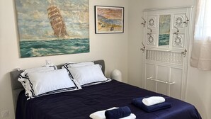 A neatly made bed with navy blue bedding, a painting of a ship on the wall, and a white door with decorative elements. - La voile Bleue - A stopover with your feet in the water (Barbâtre)