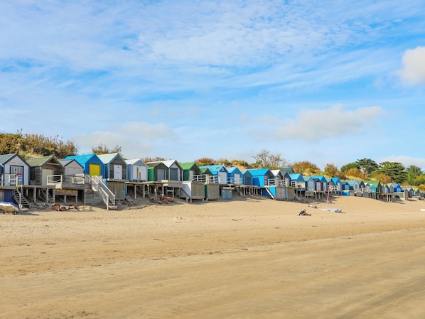 A row of beach huts with wooden staircases leading up to them, situated on a sandy beach. - Lle Te (Pwllheli)