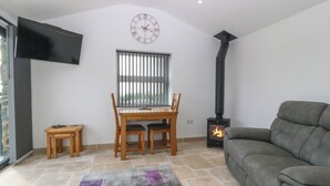 A living room with a fireplace, a dining table with chairs, a sofa, a clock on the wall, and a TV mounted on the wall. - Tyddyn Bach (Holyhead)