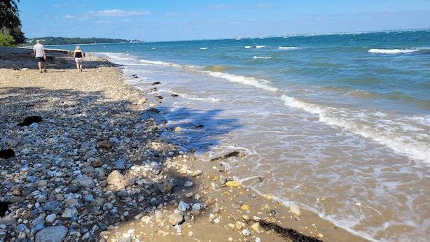 Egy kavicsos strand, ahol két ember sétál, tiszta az ég, és a tenger csendes.