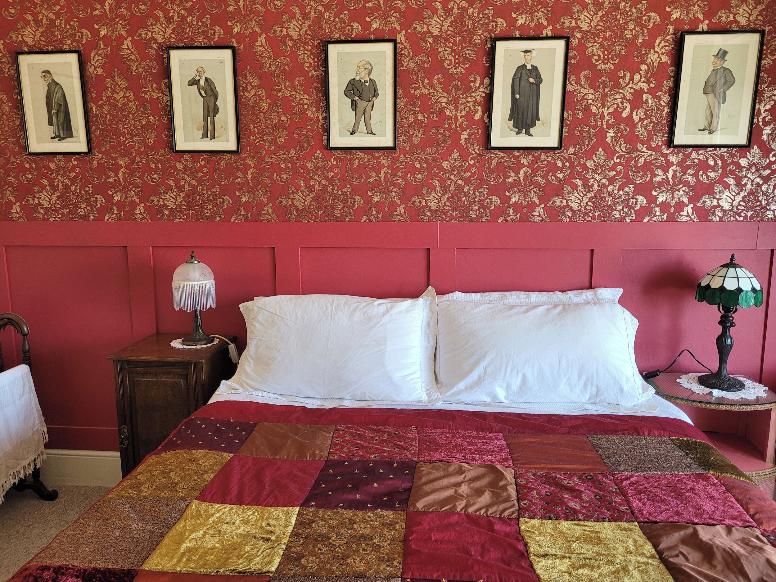 Un dormitorio con un papel tapiz rojo estampado, un edredón acolchado y cuatro cuadros en la pared.
