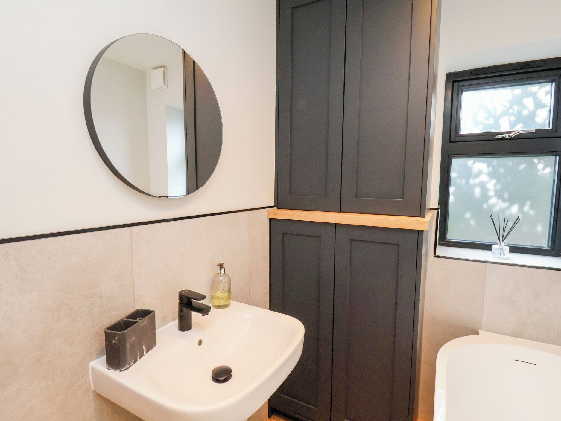A bathroom with a round mirror, a white sink, a dark wooden cabinet, and a window with a view of trees.