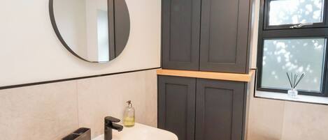 A bathroom with a round mirror, a white sink, a dark wooden cabinet, and a window with a view of trees.