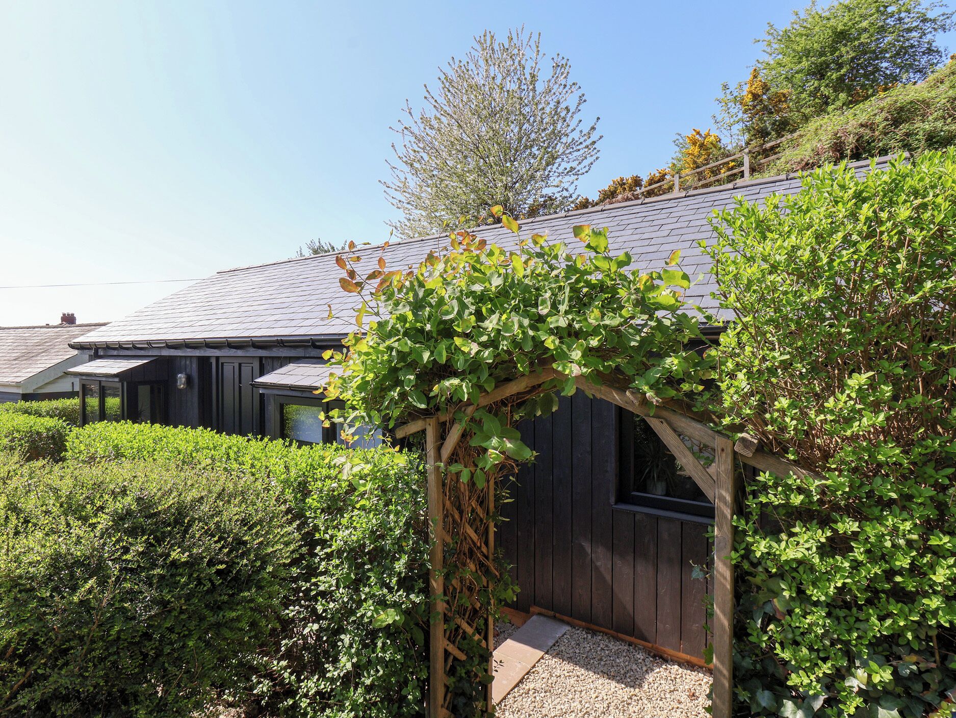 A residential property with a dark wooden exterior, a sloped roof, and a garden area with dense greenery.