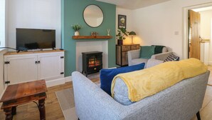 A living room with a television on a cabinet, a fireplace, a sofa with a yellow blanket, and a hanging light fixture. - Bear Cottage (Matlock)