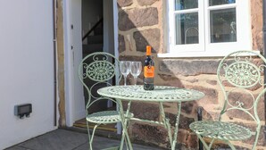 A stone building with a white door and window, a small outdoor table with a bottle of wine, and two chairs. - Bear Cottage (Matlock)