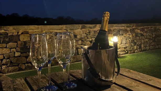 Une bouteille de champagne dans un seau en métal, posée sur une table en bois, avec des verres vides à côté, le tout sur fond d’un mur de pierre et d’une zone herbeuse.