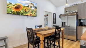 Un coin repas avec une table et des chaises en bois, un grand téléviseur fixé au mur et une cuisine équipée d’appareils électroménagers en acier inoxydable.