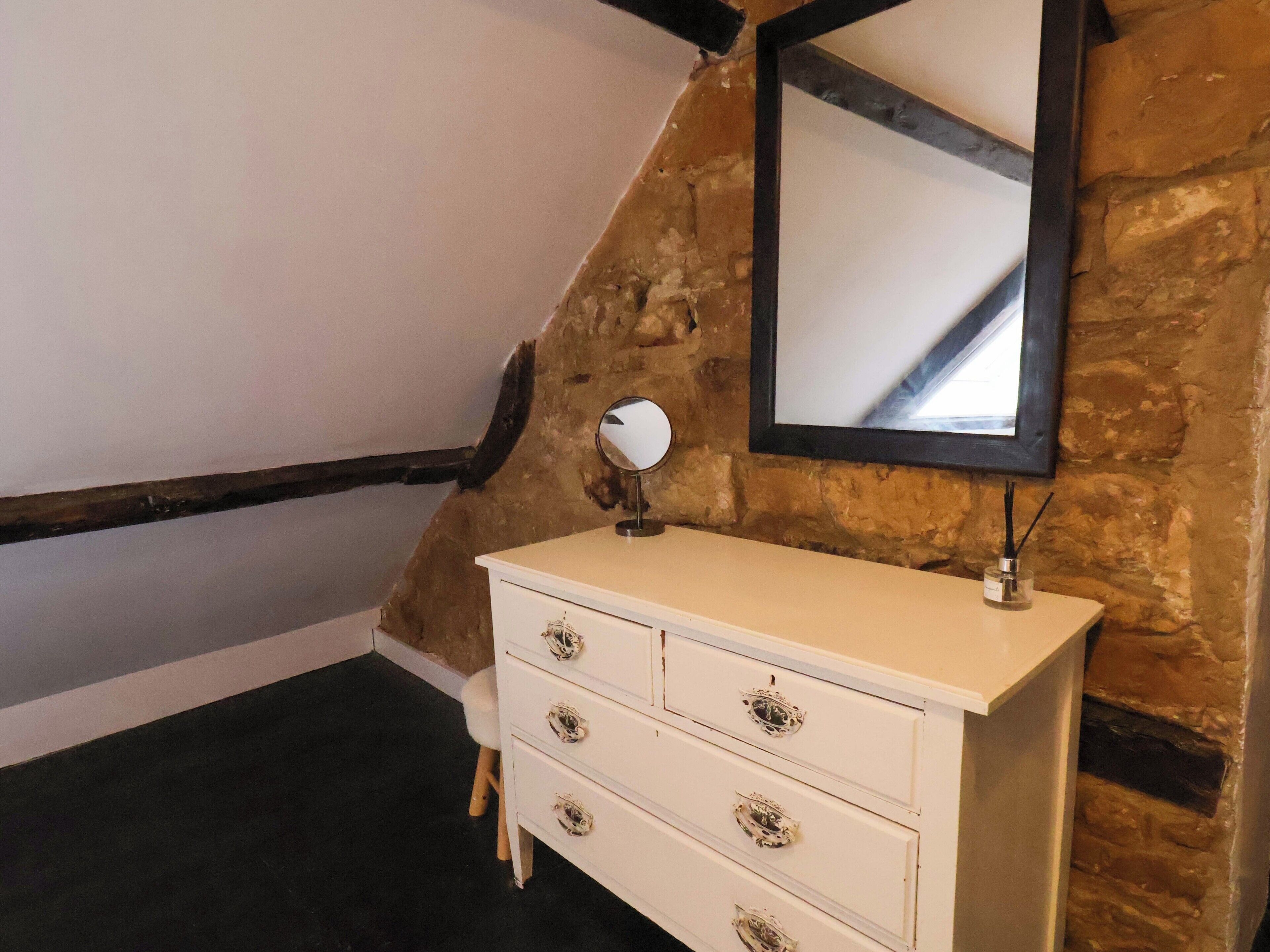 A white dresser with ornate handles, a round mirror, and a small container with reed diffusers. The room features exposed brick walls and a sloped ceiling with wooden beams.