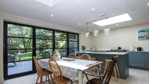 A modern kitchen with a large island, dining table with chairs, and a view of a garden through large windows. - Islwyn (Bala)