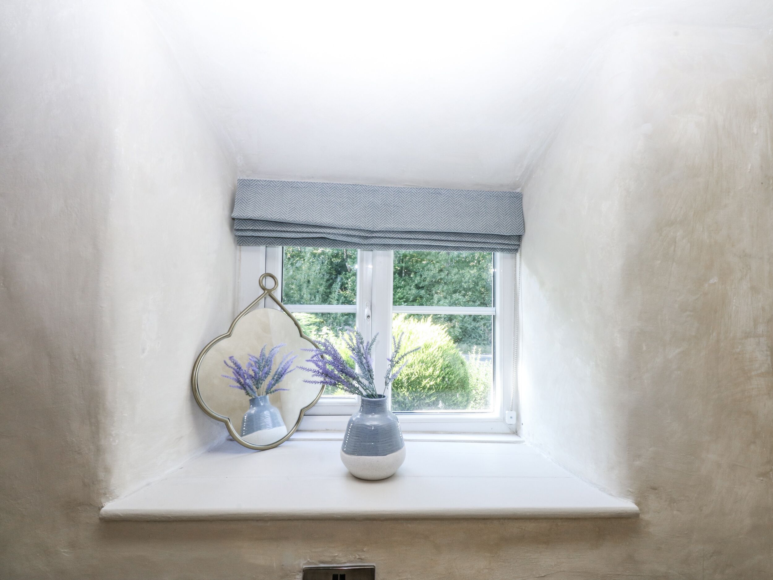 A small window with a white sill, a gray and white chevron-patterned valance, and a vase with purple flowers.