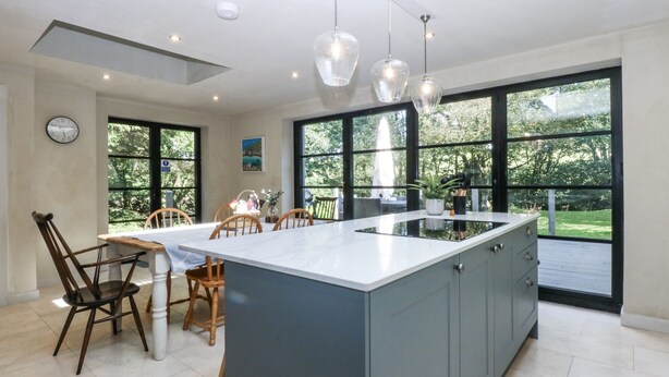 A modern kitchen with a large island, wooden chairs, and large windows offering a view of the outdoors. - Islwyn (Bala)